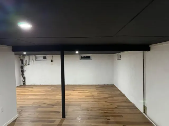 Empty basement room with white walls, wood floor, black ceiling, and a central support post