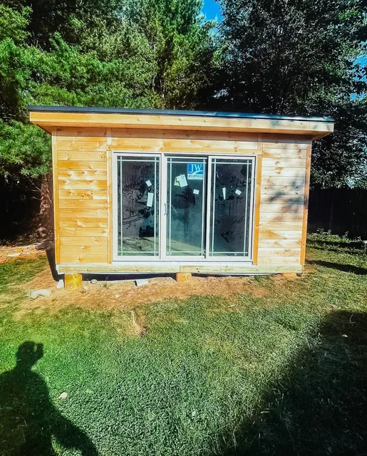 Small wooden shed with sliding glass doors in a grassy backyard, surrounded by trees and sunlight.
