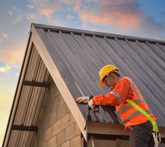 Roofer in safety gear installing metal panels on a roof against a sunset sky.