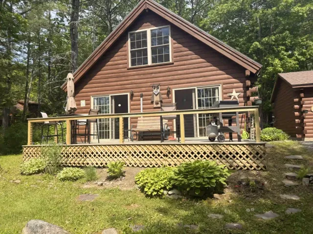 Log cabin with a deck and railing, surrounded by green trees and shrubs.