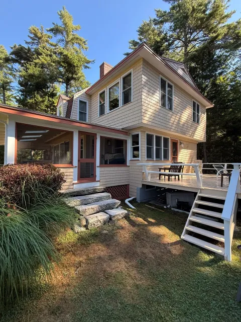 Two-story beige house with red roof, porch, and white stairs on a wooded hillside