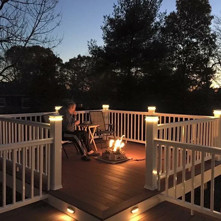 A woman sitting on a patio at sunset, drinking from a mug, with a fire pit in front and a white wooden railing surrounding the deck, illuminated by small lights on the posts.