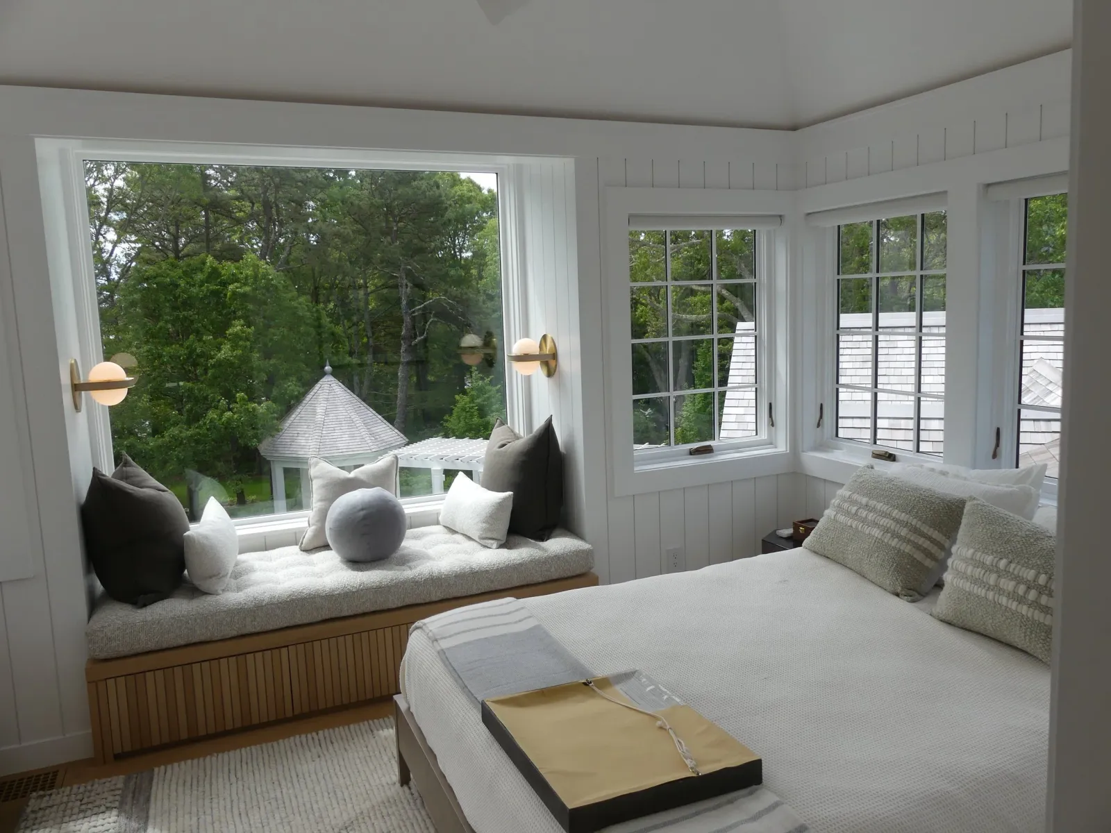 A cozy bedroom with a window seat with pillows and a view of a backyard with trees and a gazebo, and a bed with pillows and a tray on top.
