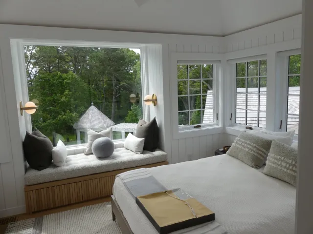 A cozy bedroom with a window seat with pillows and a view of a backyard with trees and a gazebo, and a bed with pillows and a tray on top.