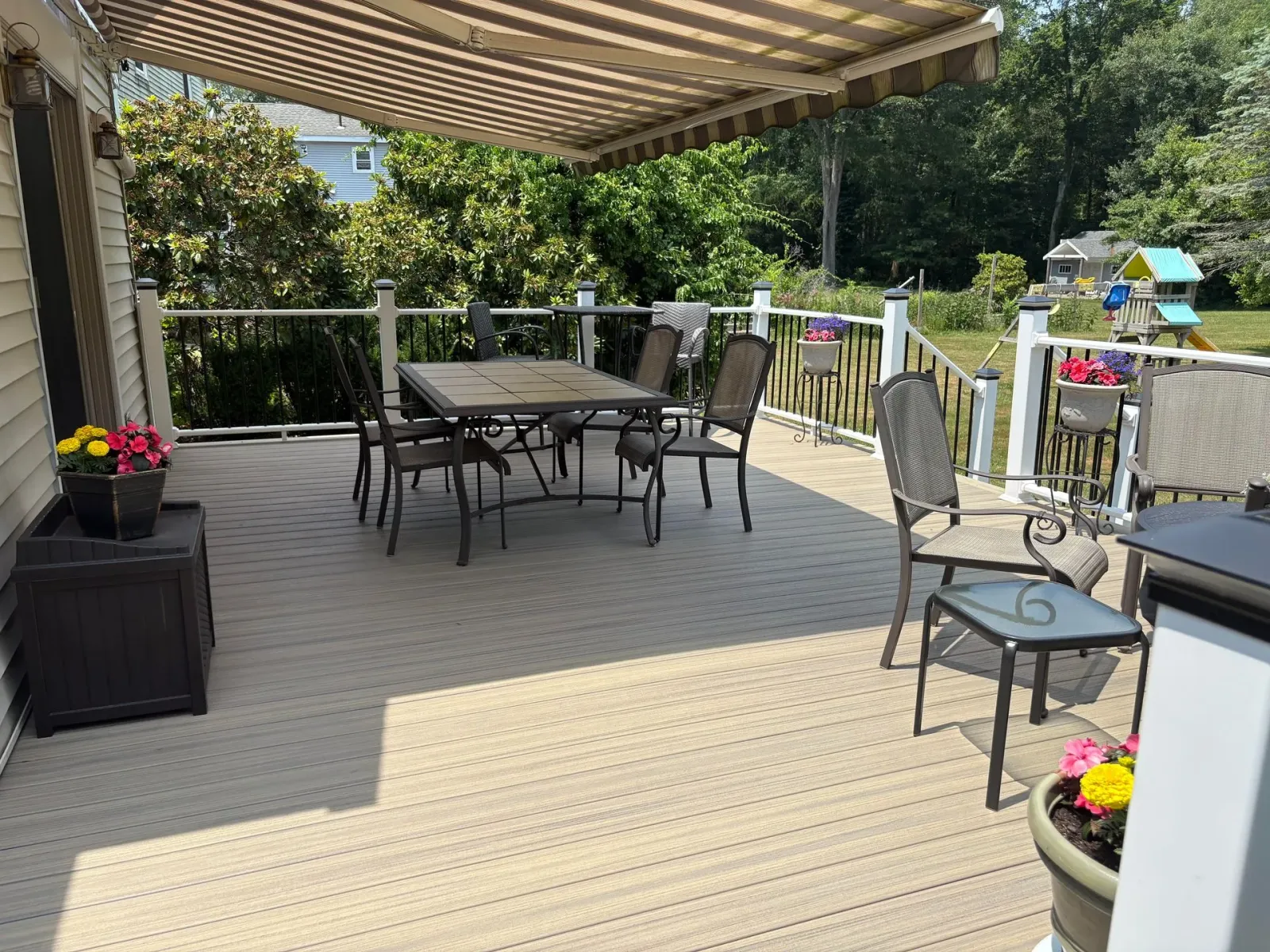 Deck with outdoor dining set under awning, potted flowers, and green yard in the background.