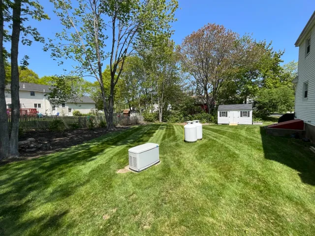 A backyard with a grassy lawn, several trees, a white shed, and two white outdoor appliances, all under a clear blue sky.