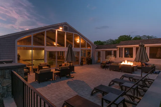 An outdoor patio with lounge chairs, umbrellas, a fire pit, and a building with large windows and a swimming pool inside at dusk.