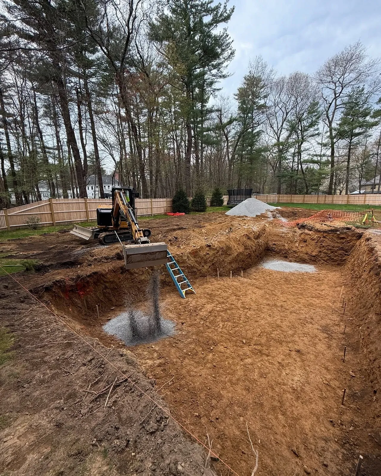 Excavated backyard with backhoe and ladder; dirt and mulch visible. Trees and fence in background.