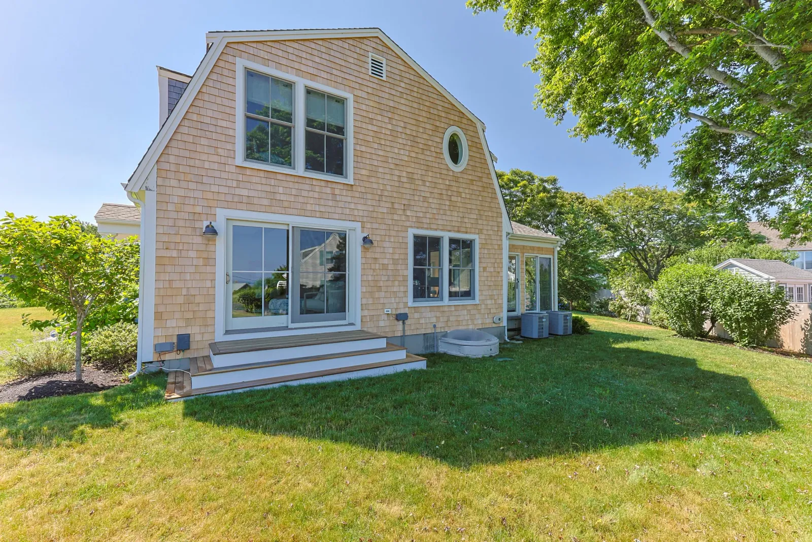Backyard of a house with beige wooden siding, sliding glass door, and stairs leading to the lawn. Green grass, trees, and neighboring houses are visible. Custom deck installation on Cape Cod by Watkins Works.