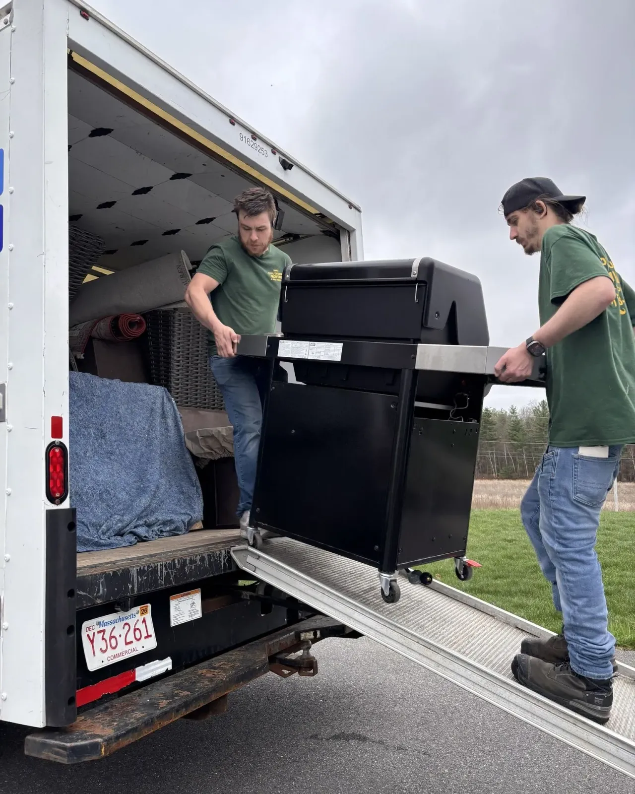 Two movers in green shirts carrying a black cabinet up a ramp into a moving truck.