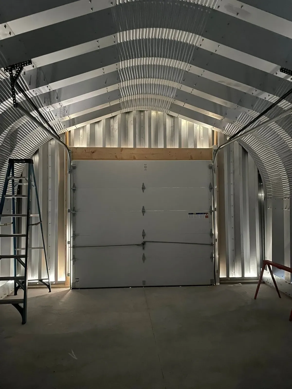 Interior of a metal building with a closed garage door, lit by overhead lights, with a ladder on the left.