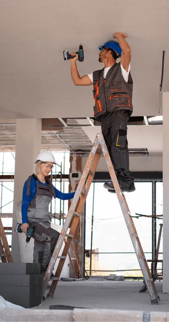 A male worker on a ladder drills into a ceiling, while a female supervisor observes in a construction site. both wear hard hats and safety vests.