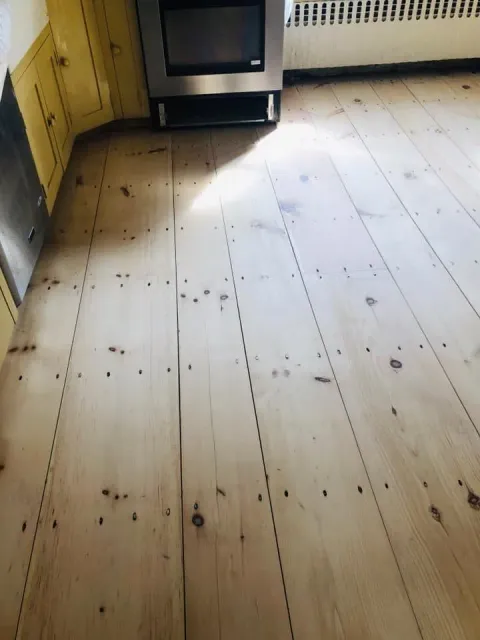Light-colored wooden floor with dark knots and small nails. Kitchen setting with an oven and yellow cabinets.