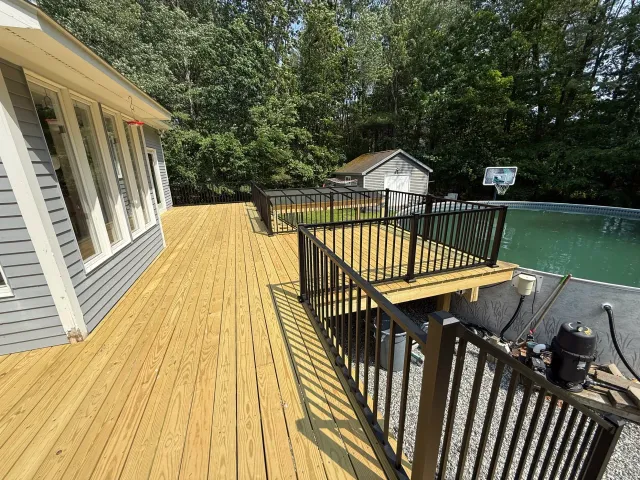 Newly built wooden deck with black metal railings, adjacent to a house with gray siding, overlooking a backyard with a fenced pool and a basketball hoop, surrounded by trees.