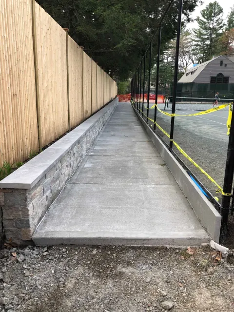 A sidewalk bordered by a stone wall and fence leads through a construction site with yellow caution tape.