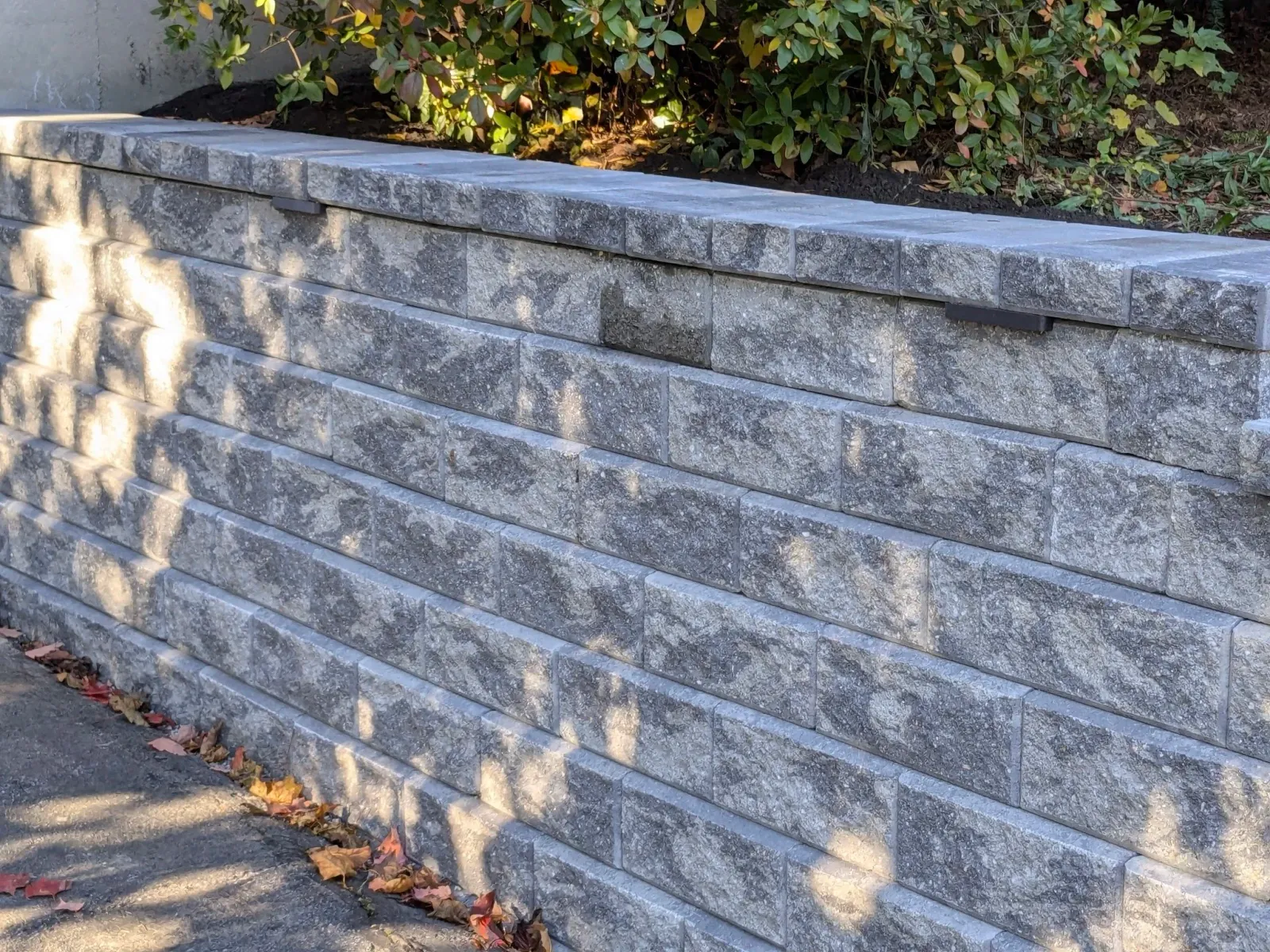A stone retaining wall with rectangular blocks, with some fallen autumn leaves along the base and bushes behind it.
