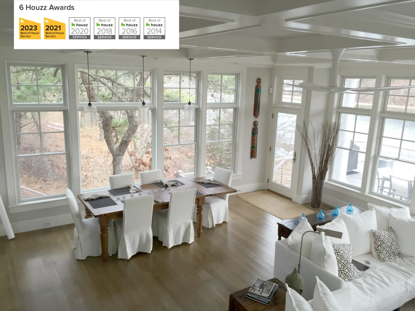 Bright living and dining room with large windows, white furniture, a wooden table, and decorative items, featuring a view of trees and a body of water outside.