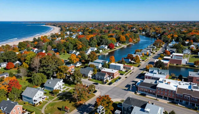 Aerial view of a coastal town with a beach, river, roads, and autumn-colored trees.