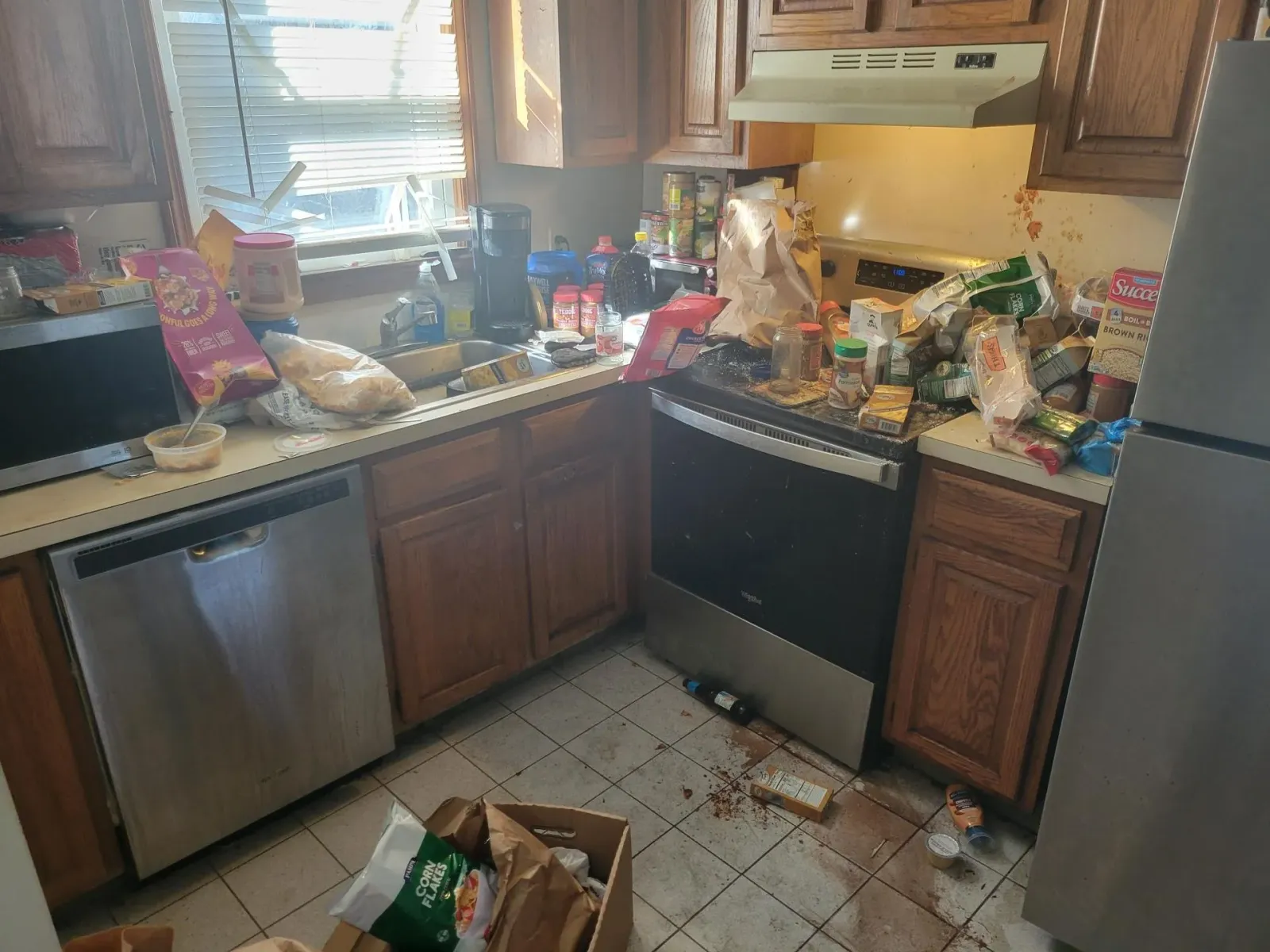 Messy kitchen with counters cluttered with food items, appliances, and spilled liquid on the floor.