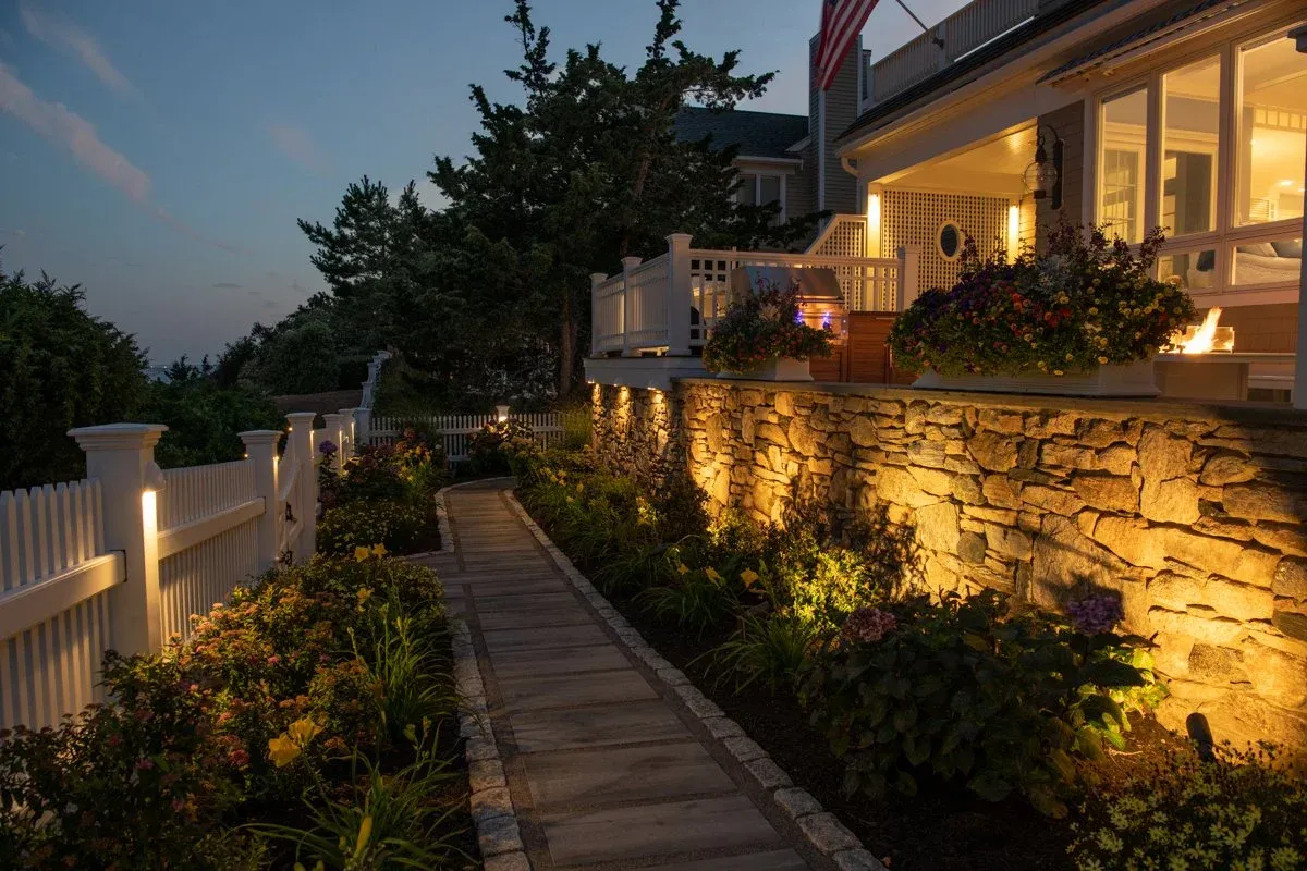 A backyard garden pathway illuminated by landscape lighting along a stone retaining wall, with a house featuring a screened porch and flower planters at dusk.