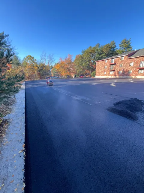 Newly paved asphalt parking lot with a clear blue sky, buildings, and fall foliage in the background.