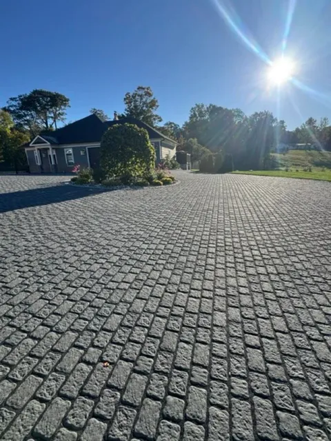 A paved driveway in front of a house on a sunny day, with trees and greenery in the background.