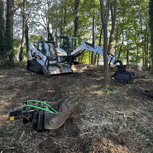 Bobcat excavator in a wooded area with attachments, clearing debris.