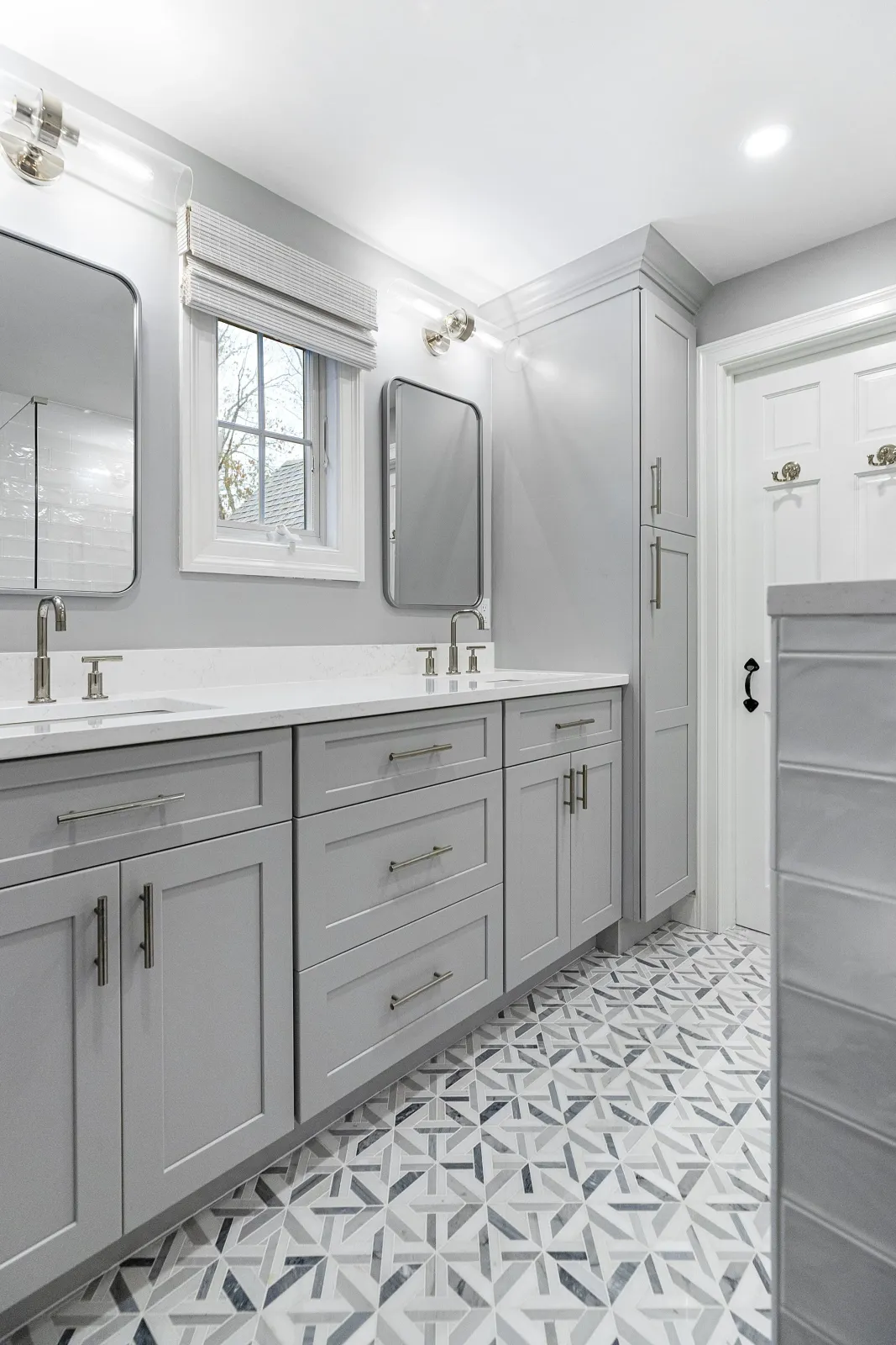A modern bathroom with a double vanity, gray cabinets, white countertops, two mirrors, and a small window. The floor has geometric patterned tiles.