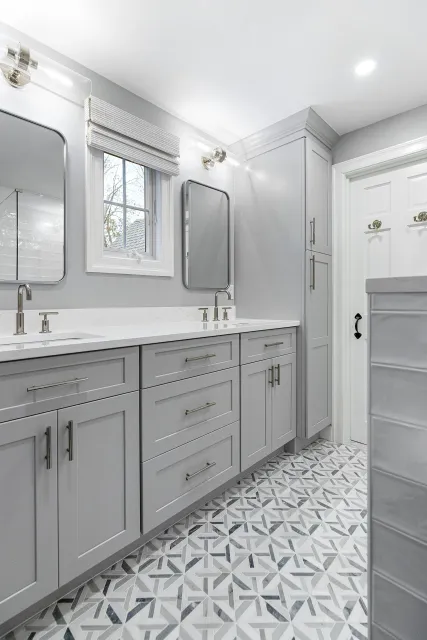 A modern bathroom with a double vanity, gray cabinets, white countertops, two mirrors, and a small window. The floor has geometric patterned tiles.