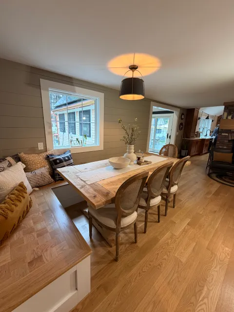 A cozy dining and living area with wooden flooring, a large wooden dining table, rattan chairs, a corner with a couch decorated with patterned cushions, a window letting in natural light, and a view of a porch and other houses outside.