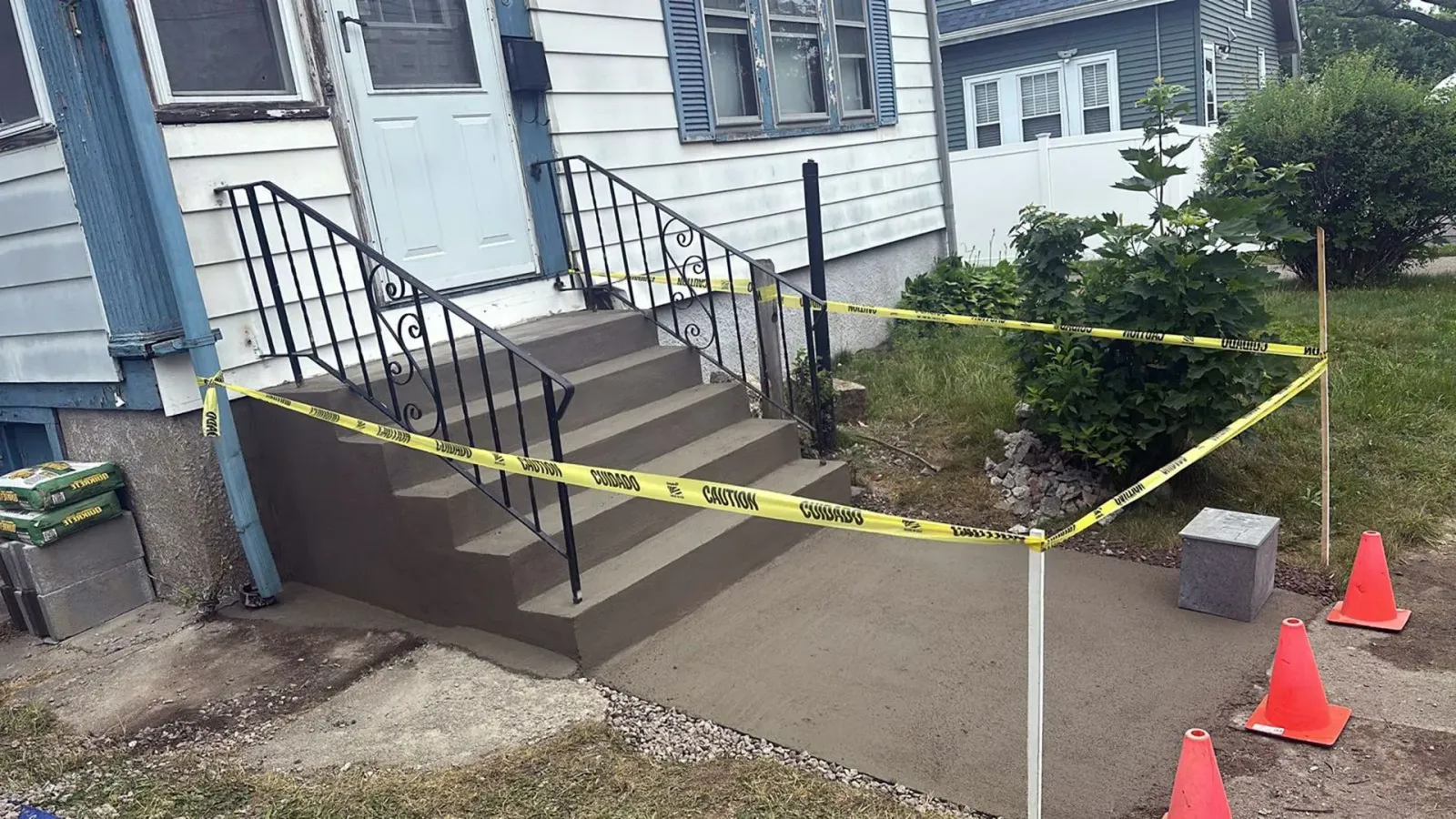 Front porch under construction, with taped-off walkway, black railings, and orange cones beside a house.