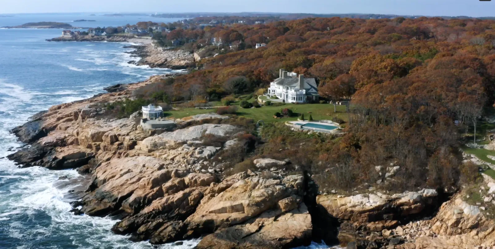 A coastal landscape with rocky cliffs, ocean waves, and large houses surrounded by trees with autumn foliage.