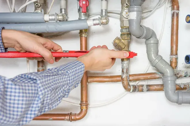 A person in a blue-checkered shirt uses a red wrench to tighten a brass fitting on a copper pipe plumbing system.