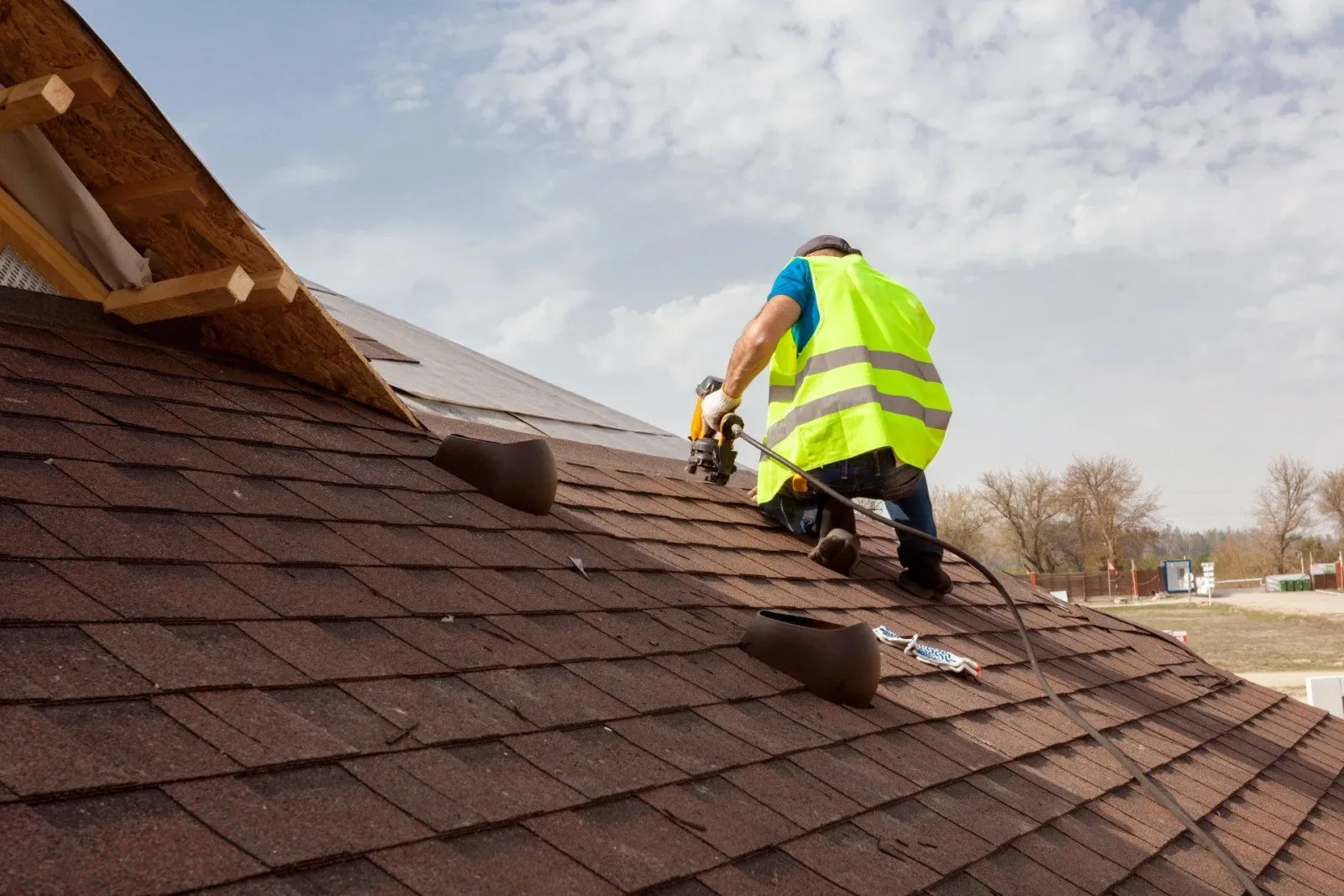 Roofer in safety vest using a nail gun on a shingled roof, blue sky background.