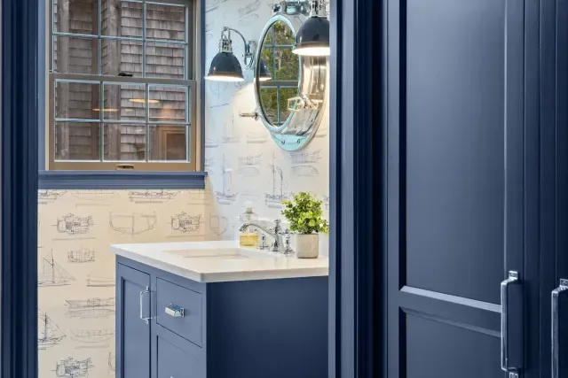 Blue bathroom vanity with marble countertop, round mirror, window, and wall sconces.