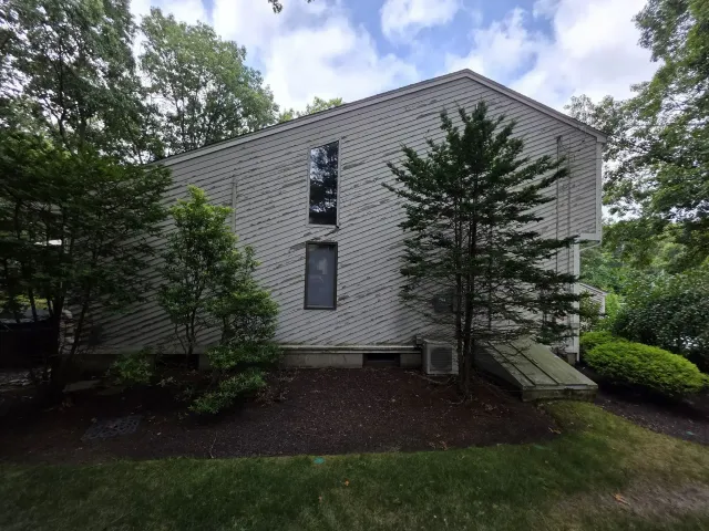 Two-story white brick house with a small ramp, trees, and shrubs in a wooded yard.