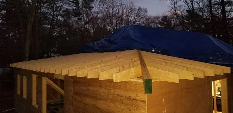 A wooden structure with an unfinished roof. A blue tarp covers a portion of the roof. Dark trees in the background.