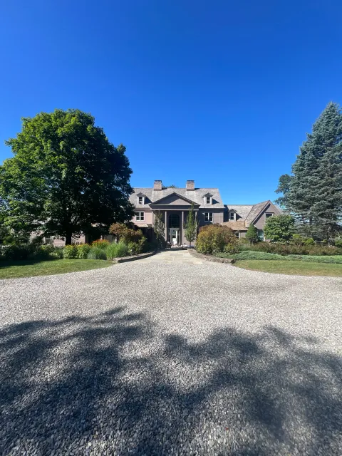 Large gray house with a stone pathway leading to the front entrance, surrounded by green trees and bushes under a clear blue sky.