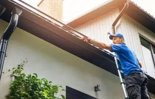 Man on ladder cleaning house gutters as part of exterior cleaning. He wears a blue shirt and cap, focused on the task. The house has white siding and dark trim.