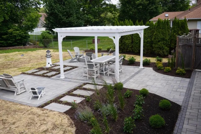 A backyard with a paved patio area featuring outdoor furniture including a dining table with chairs under a white pergola. There are Adirondack chairs, a small lighthouse decoration, and landscaped garden beds with plants and bushes. A chain-link fen