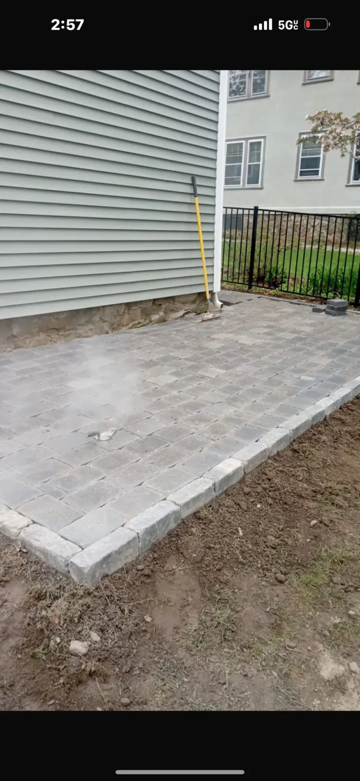 A newly constructed gray paver patio next to a house with light green siding and a small garden.