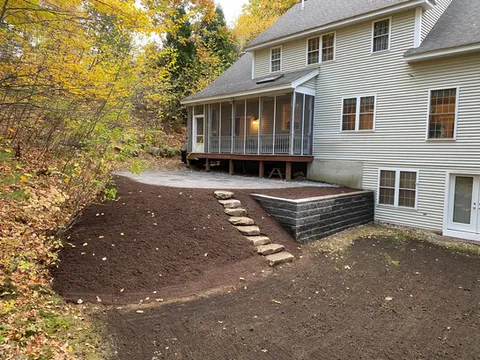 A newly graded backyard slopes gently away from a large gray house with an elevated wooden deck and warm exterior lighting. Freshly spread soil covers the yard, while a set of stone steps leads up to the deck. Tall trees with autumn foliage border the property, giving the outdoor space a secluded woodland feel.