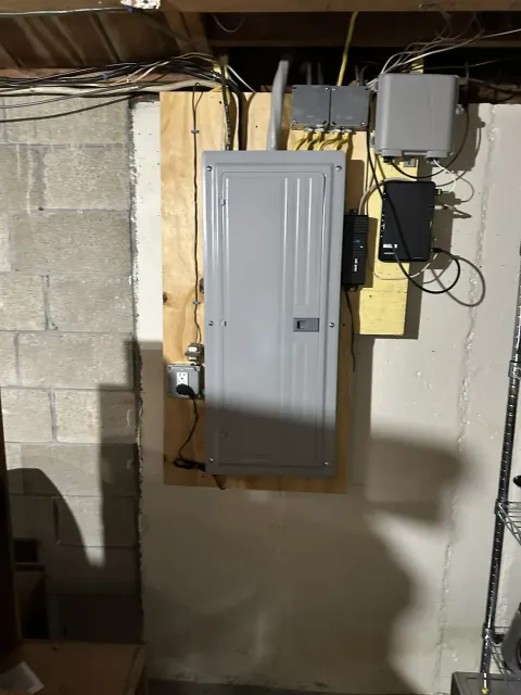 Electrical panel mounted on a plywood backboard in a basement with circuit wiring run overhead