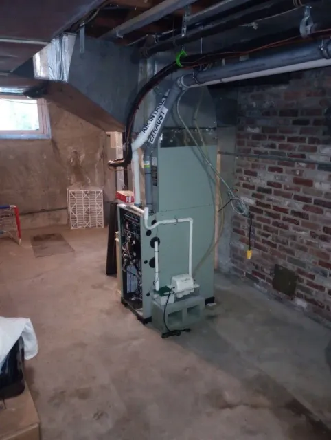 Furnace in a basement; gray metal unit against brick wall, with pipes and ductwork.