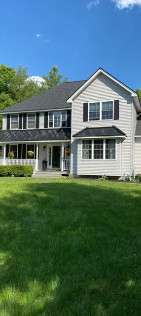 A two-story traditional home with light gray siding, black shutters, a wrap-around porch, and a lush green lawn.