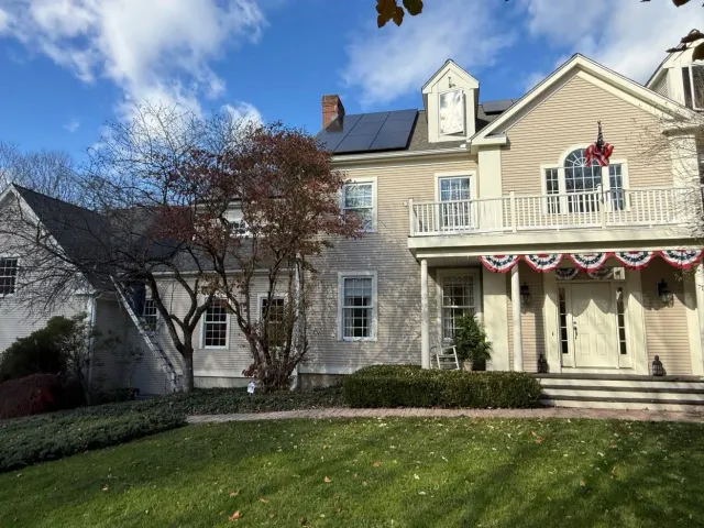 A beige two-story house with a wooden porch, flag decorations, solar panels on the roof, and a front lawn under a blue sky.