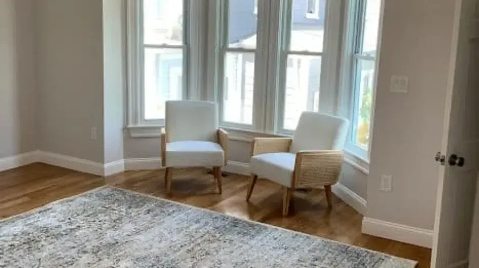 Bright living room with bay windows, two accent chairs, hardwood floors, and area rug, located in the Greater Boston area