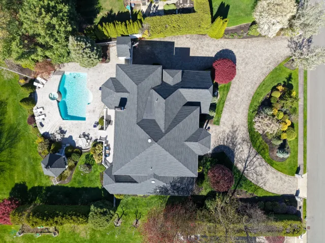 Aerial view of a large house with a circular driveway, landscaped garden, and a backyard pool surrounded by trees, shrubs, and green lawns in a suburban neighborhood.