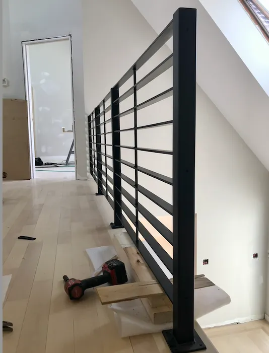 A black metal railing installed along the edge of a staircase in a room under renovation, with a cordless drill and wooden pieces on the floor nearby.