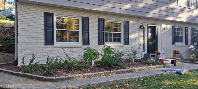 Single-story white brick house with black shutters, front door, and a small landscaped yard in autumn.